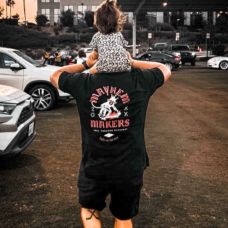 A man, exuding agility, moves through a parking lot with a child joyfully perched on his shoulders and wearing Mayhem Makers - Black.