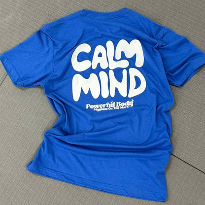 A premium cotton t-shirt in royal blue lies on the floor, showcasing "Calm Mind, Powerful Body" in bold white letters, perfect for promoting mindfulness.