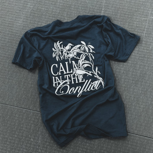 The Calm In The Conflict - Indigo Navy T-shirt, made from premium cotton, rests on a textured background, featuring white text and a bird illustration with the words "Calm in the Conflict." This piece from the "Serenity with Style" collection embodies both elegance and comfort.