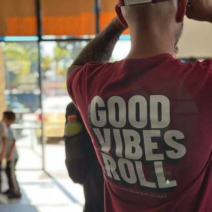 A person in a cardinal red shirt from the "Good Vibes Roll" collection is standing indoors near a window, embodying the camaraderie found in Jiu Jitsu.