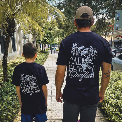 A man and a child stroll down the sidewalk, both sporting matching "Calm In The Conflict - Black" t-shirts. Their peaceful design stands out amidst the trees and buildings, crafted from premium cotton to symbolize peace and unity in style.