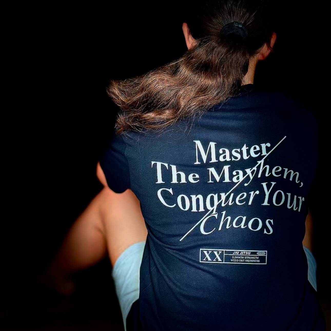 The person sits with their back to the camera, wearing the "Master The Mayhem / Conquer Your Chaos - Black" shirt, which features bold white text on a dark background.