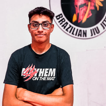 A person with glasses stands confidently, arms crossed, wearing a "Clawed Mayhem - Black" shirt. They pose proudly in front of a Brazilian Jiu Jitsu emblem, embodying the spirit of their sport and celebrating their journey on the mat.