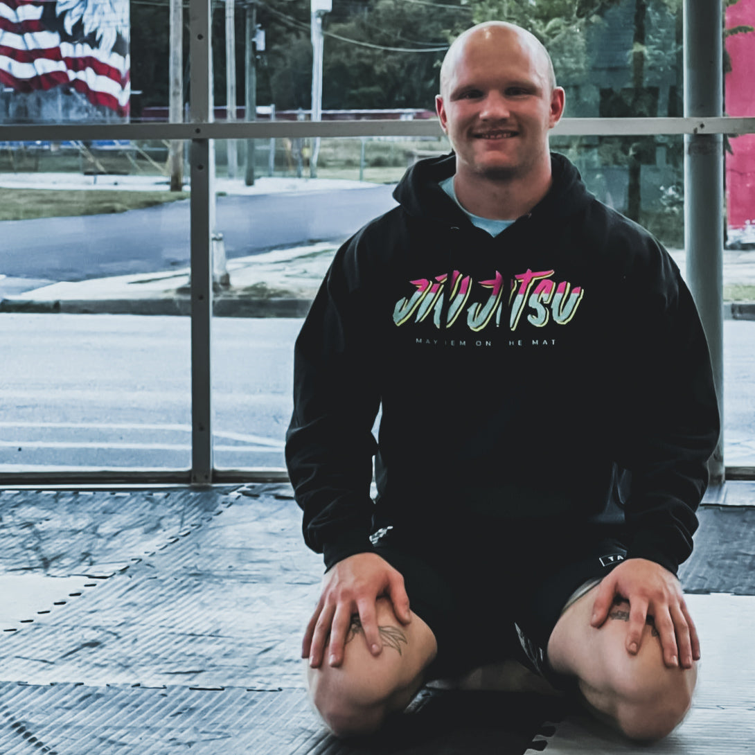 A person wearing a "City Connect - Black" hoodie kneels on a gym mat in a room with large windows, embodying the spirit of camaraderie that thrives within this close-knit community.