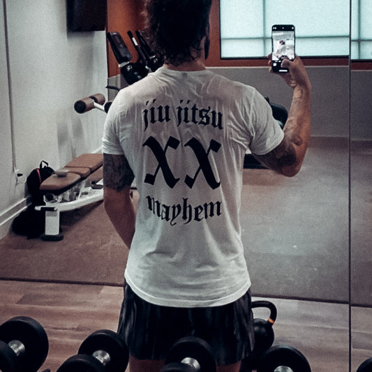 In the gym, a person snaps a selfie wearing a white "Team Mayhem" t-shirt, with dumbbells scattered and gym equipment in the background, proudly showcasing their apparel.