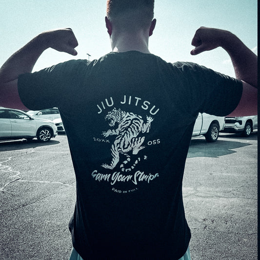 In a parking lot, someone flexes in a black "Earn Your Stripes" T-shirt, showcasing a bold tiger design with "Jiu Jitsu, Earn Your Stripes," embodying strength and dedication.