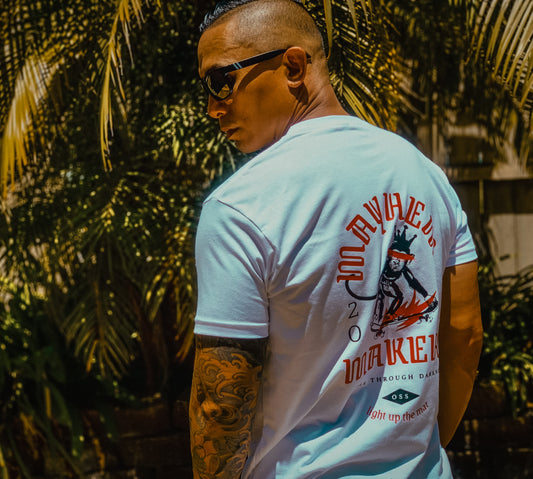 A person wearing sunglasses and a "Mayhem Makers - White" graphic t-shirt with a tattooed arm stands outdoors, exuding an air of inner strength.