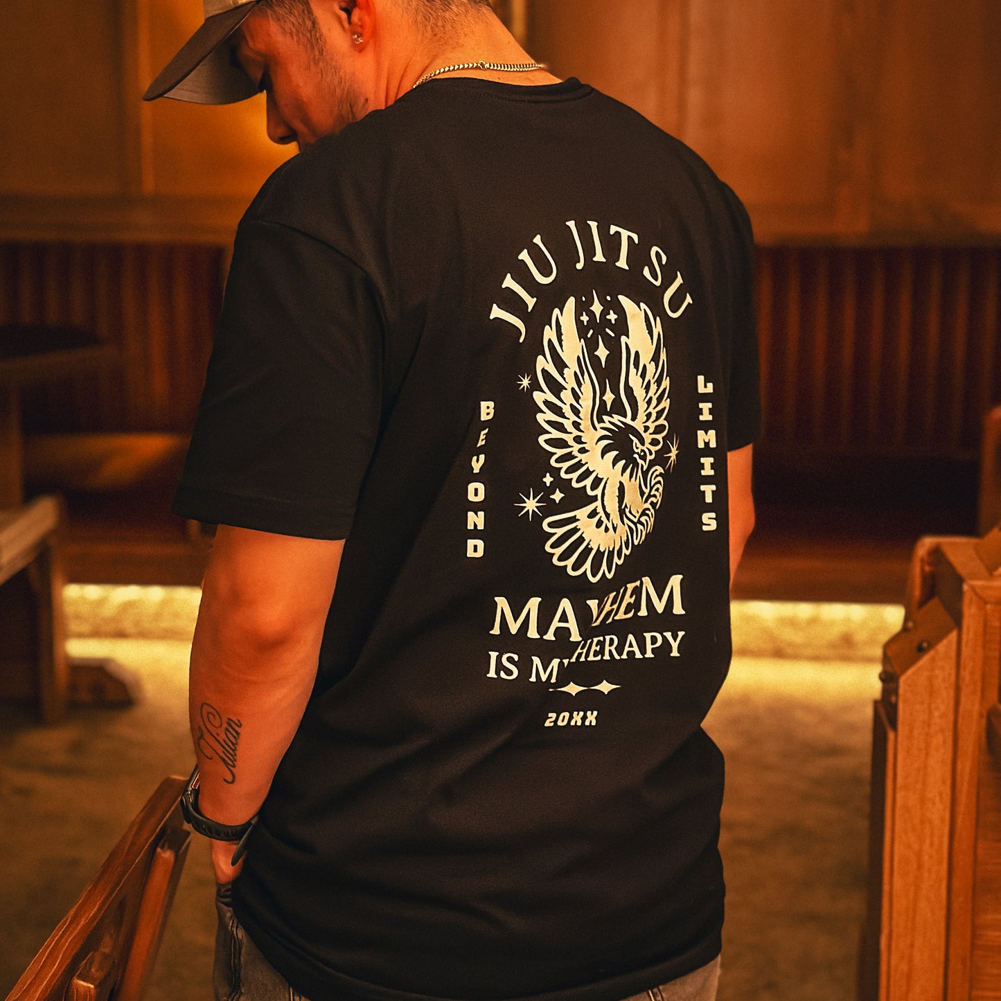 A man stands indoors wearing the "Beyond Limits - Black" T-shirt, featuring an eagle graphic and the phrase "Jiu Jitsu Beyond Limits, Mayhem is My Therapy.