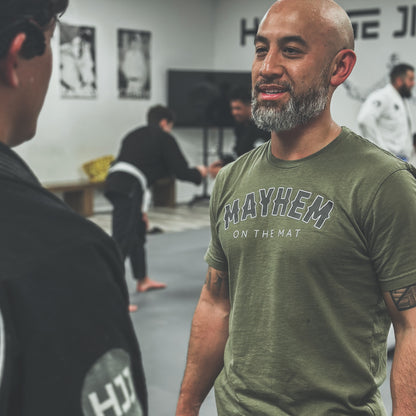 A person wearing a Military Green shirt with the Rocker logo smiles in conversation during a martial arts class, while others practice Jiu Jitsu in the background, all mastering the mayhem together.