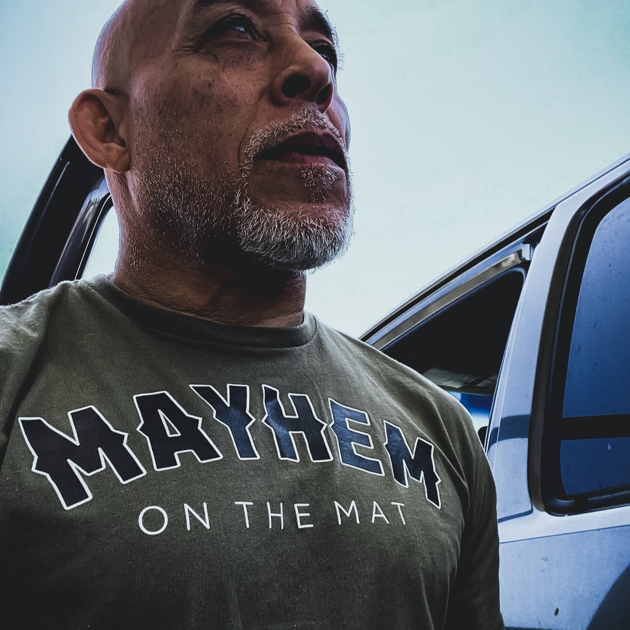 A man with a beard, wearing a shirt that reads "MAYHEM ON THE MAT" paired with the discreet Rocker logo in military green, stands next to an open car door.