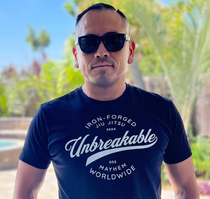 A person wearing sunglasses stands confidently outdoors, embodying resilience in the "Unbreakable - Black" t-shirt. The blurred background suggests trees and a small pool, enhancing the serene ambiance.