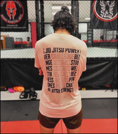 With long hair tied up and wearing the Strength & Power - White shirt, the person stands in the MMA gym, exuding an aura of empowerment and strength as they face away from onlookers.