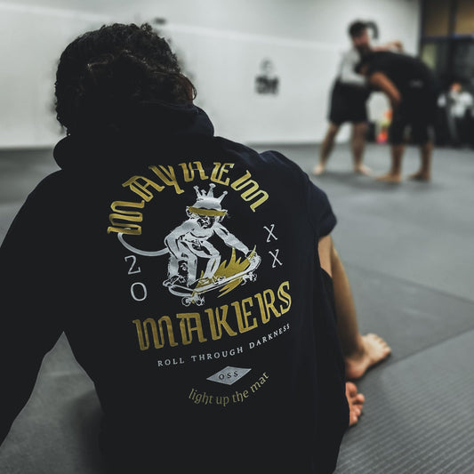 A person sits confidently on a gym mat, wearing a "Mayhem Makers - Navy" shirt emblazoned with "MAKERS," embodying the essence of strength. In the background, two individuals gracefully practice Jiu Jitsu, their movements precise and harmonious.