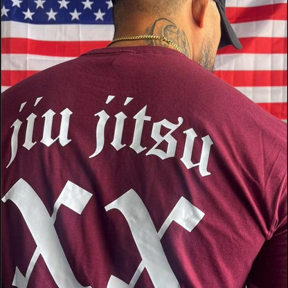 A person proudly wears a maroon Team Mayhem shirt with "Jiu Jitsu XX" prominently displayed on the back, standing confidently in front of an American flag.