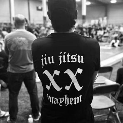 A person sporting a "Team Mayhem - Black" shirt stands indoors, fully embodying the Jiu Jitsu lifestyle as they face an exhilarating event.