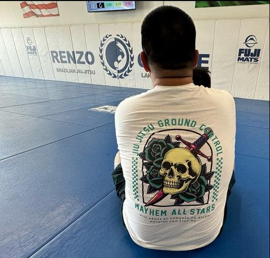 Dressed in the "Jiu Jitsu Ground Control - White" t-shirt featuring a striking skull and rose graphic, an individual sits on a blue martial arts mat, embodying true mastery. With their back turned to the Brazilian Jiu-Jitsu logos that adorn the walls, they reflect a deep commitment to overcoming challenges.