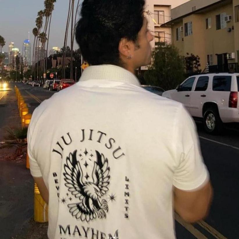 A person stands on a palm-lined street at dusk, wearing the "Beyond Limits - White" shirt, featuring an eagle design and "Jiu Jitsu Mayhem" on the back, symbolizing the spirit of overcoming challenges.