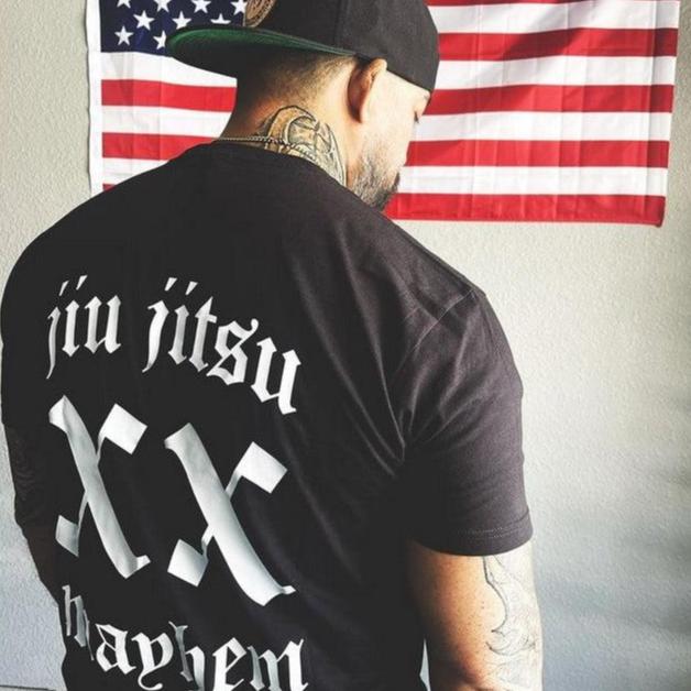 A man with tattoos, proudly wearing his "Team Mayhem - Black" shirt and cap, embodies the Jiu Jitsu lifestyle as he stands confidently before an American flag.