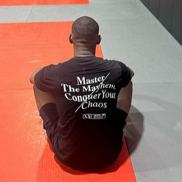 A person sits on a red and gray mat in the gym, facing away, with punching bags hanging in the background. The back of their shirt boldly displays, "Master The Mayhem / Conquer Your Chaos - Black," mirroring their determination to tackle any challenge that comes their way.